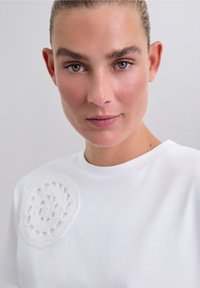 Femme à la peau claire et aux yeux verts regardant vers l'avant, portant un haut blanc avec un détail en dentelle circulaire sur l'épaule, sur un fond uni.