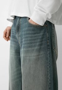 Vaaleat siniset farkut, joissa on rento istuvuus, etutaskut, kontrastivärinen tikkaus ja sileä pinta. Vyötärönauha näkyy hieman.