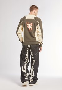 Felpa grigia e crema con grafica arancione, abbinata a jeans neri a gamba larga con design di testo bianco. Sneakers bianche completano il look.