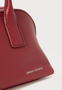 Punainen nahkainen käsilaukku nurkassa, jossa on ommeltu kahva ja valkoinen Armani Exchange -logo sileällä tekstuuripinnalla valkoista taustaa vasten.