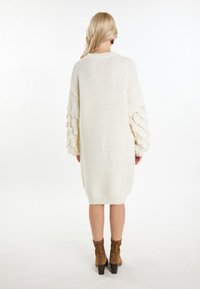 Abito in maglia sovradimensionato color crema con maniche a palloncino testurizzate e polsini a costine. Abbinato a stivali alla caviglia in suede marrone e uno sfondo neutro.
