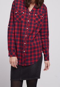 Rood en marineblauw geruit shirt met lange mouwen, twee borstzakken en witte knopen. Gecombineerd met een zwarte denim rok.