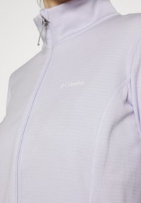 Giacca a zip di colore viola chiaro realizzata in tessuto texturizzato, con colletto alto e un piccolo logo bianco sul davanti.