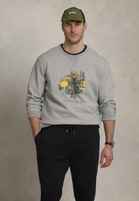 Sweatshirt cinza com um gráfico de um urso em traje militar ao lado de um veículo amarelo, combinado com calças de moletom pretas e um boné verde.