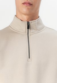 Beige half-zip pullover made of soft fabric. Features a high collar and a contrasting zipper detail, with a smooth and slightly textured surface.