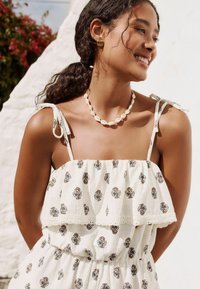 Witte jurk met zwarte bloempatronen, ruches bij de halslijn, dunne bandjes, schelpketting, gestructureerde stof. Model met donker, krullend haar.