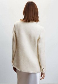 Cremefarbener Tweed-Blazer mit strukturiertem Finish, ausgefranstem Saum und goldenen Knopfakzenten an den Ärmeln. Rückansicht dargestellt.