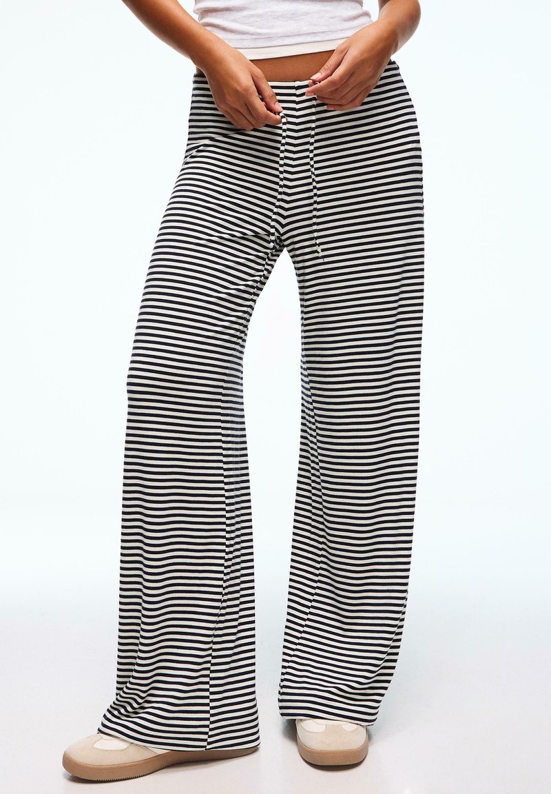 Personne portant un pantalon large à rayures horizontales noires et blanches, ajustant le cordon de serrage à la taille, assorti à des baskets beiges.