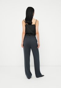 Frau mit schwarzen Haaren, die ein schwarzes ärmelloses Oberteil und eine dunkle, gestreifte, weit geschnittene Hose trägt, steht vor einer weißen Wand.