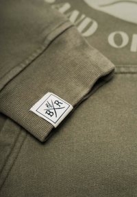 Olivgrön sweatshirt med ribbade ärmslut, med en vit etikett med "B/R"-logotypen. Tyget har ett texturerat, vävt utseende.