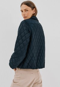 Quilted marineblå jakke med krave, der fremviser en diamantmønstret tekstur og en kortere pasform. Modellen står med hænderne i lommerne iført beige bukser.