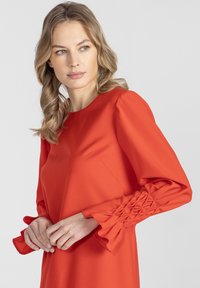 Czerwona sukienka z długimi, marszczonymi rękawami ozdobionymi fakturowanymi akcentami na nadgarstkach. Materiał wydaje się gładki, o strukturalnym kroju i okrągłym dekolcie.