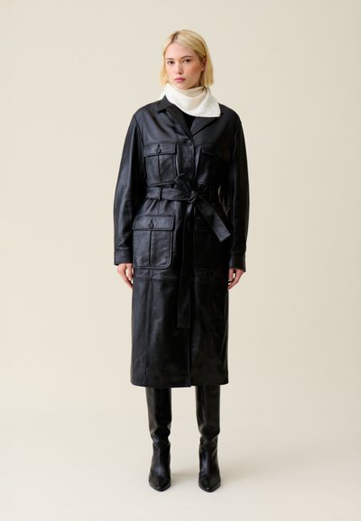 Femme portant un manteau en cuir noir ceinturé avec quatre poches à boutons, des bottes noires montantes jusqu'aux genoux, et une écharpe blanche sur un fond uni.