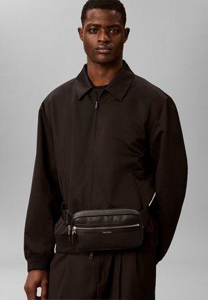 Mann trägt eine schwarze Jacke mit Reißverschluss und eine schwarze Umhängetasche mit zwei silbernen Reißverschlüssen vor einfachem grauem Hintergrund.