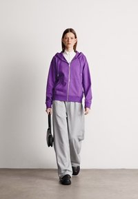 Fialová mikina so zipsom a šnúrkami, sivé voľné nohavice a čierne nazúvacie topánky. Model drží malú čiernu kabelku. Jednoduché pozadie.