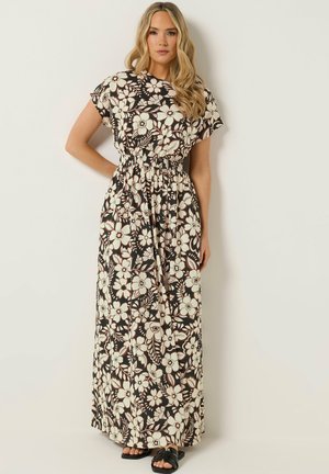 Frau mit langen blonden Haaren, die ein schwarz-weißes, geblümtes Maxikleid mit kurzen Ärmeln und elastischem Bund trägt und vor einer schlichten Wand steht.