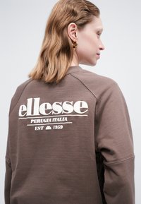 Osoba s hnedými vlasmi po pleciach, oblečená v hnedé mikine ellesse s bielym logom na chrbte, otočená doprava.