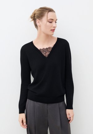 Femme aux cheveux blonds en chignon, portant un pull noir à col en V sur un top en dentelle et un pantalon plissé gris, regardant vers sa droite.