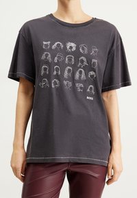 Tmavě šedé oversized tričko s bílými liniovými kresbami různých účesů. Navrženo s kulatým výstřihem a kontrastními švy.