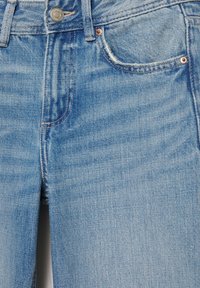 Jean en denim bleu clair en gros plan montrant le bouton avant, la fermeture éclair, les passants de ceinture et la poche avant avec des rivets en cuivre.