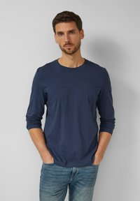 Mann mit kurzem braunem Haar und Bart, trägt ein navyblaues Langarmshirt und blaue Jeans, steht mit den Händen in den Taschen vor einer schlichten grauen Wand.