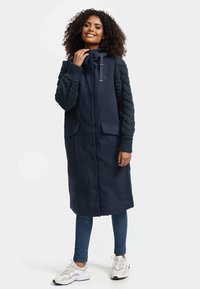 Lange marineblauwe jas met gewatteerde gebreide mouwen, voorzien van een ritssluiting, capuchon en twee voorzakken. Draagt over jeans en beige sneakers.