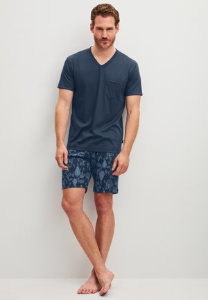 Man wearing a dark blue V-neck T-shirt with pocket and patterned navy blue shorts standing barefoot against a white wall.