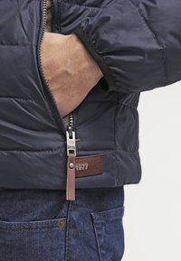 Mano nella tasca di un giubbotto trapuntato blu scuro con zip metallica e tirante in pelle, sopra la tasca dei jeans blu denim.