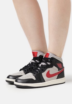Jambes portant des baskets montantes Air Jordan 1 en noir, gris et rouge avec des lacets noirs sur un fond blanc.