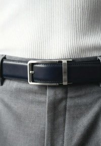 Ceinture en cuir bleu marine avec une boucle en métal argenté, présentant des détails de couture. Portée sur un pantalon en tissu gris, mettant en valeur un design épuré.