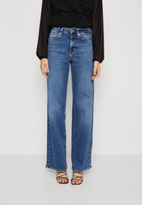 Personne portant un jean bleu taille haute à jambes larges, un haut noir transparent à manches longues et des sandales noires à brides sur un fond uni.
