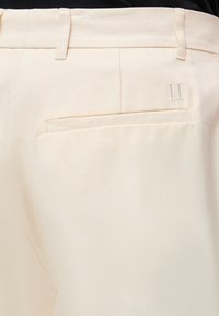 Gros plan sur la poche arrière d’un pantalon couleur crème avec passants de ceinture et un discret détail brodé du chiffre romain II au-dessus de la poche.