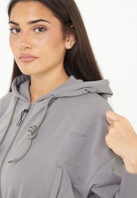 Felpa grigia con zip e cappuccio regolabile, caratterizzata da un logo sul lato sinistro del petto e un tessuto testurizzato. Il modello tiene la zip con una mano.
