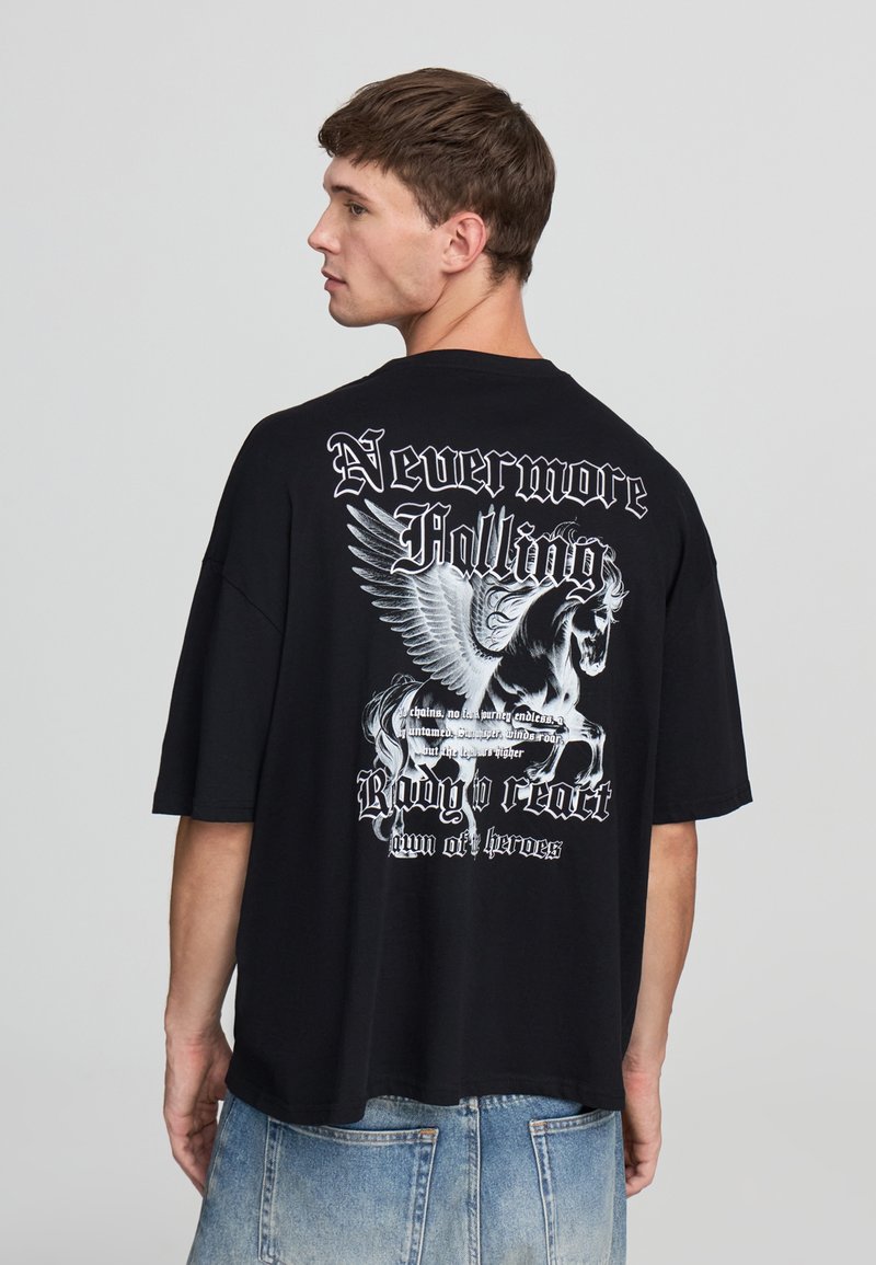 Młody mężczyzna z krótkimi brązowymi włosami nosi czarną, za dużą koszulkę z białym skrzydlatym koniem i gotyckim tekstem na plecach, w połączeniu z niebieskimi dżinsami.