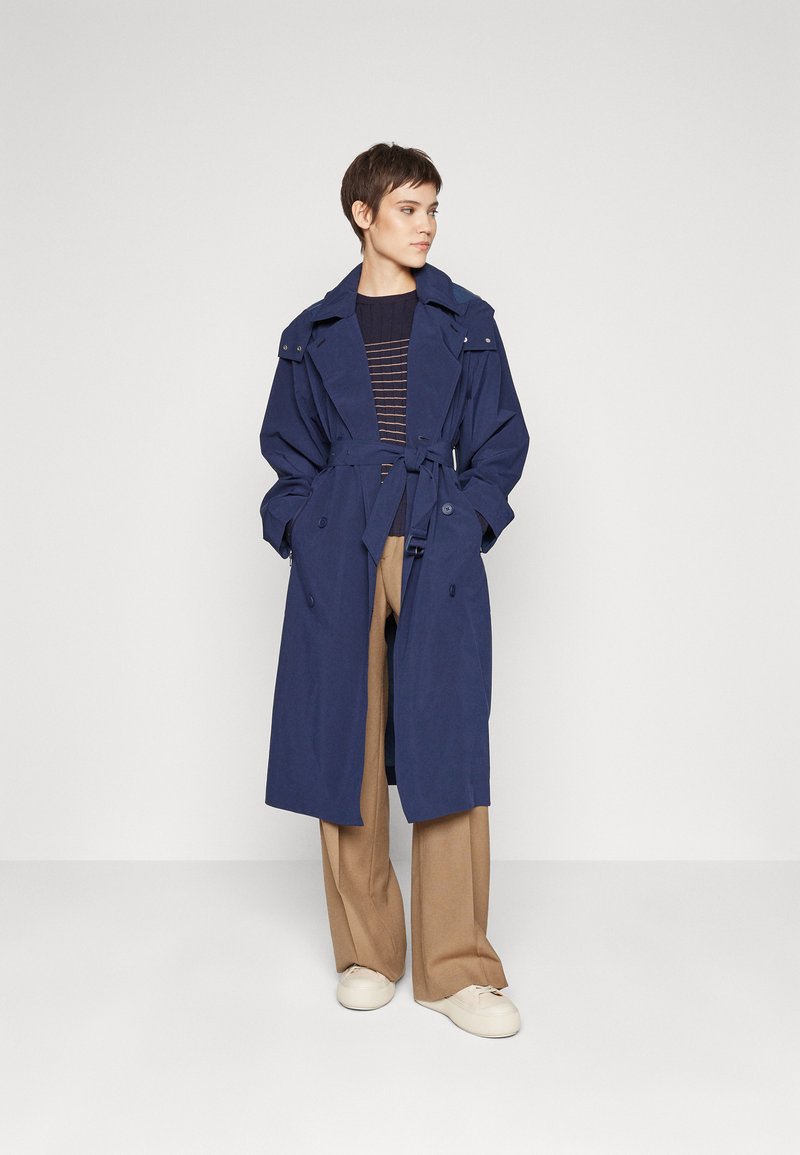 Manteau trench bleu marine avec une taille ceinturée, des boutons croisés et des épaulettes. Porté par-dessus un pull rayé et un pantalon large beige.