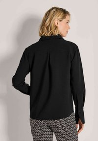Frau mit schulterlangem blonden Haar trägt ein schwarzes Langarmshirt und schwarze Hose mit weißem Muster, steht mit dem Rücken zu einem einfarbigen Hintergrund.