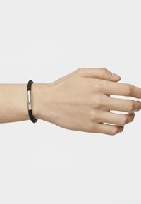 Pulsera de cuero trenzado negro con un cierre cilíndrico de plata, llevada en una mano. La pulsera presenta una textura suave y un diseño sencillo.