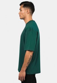 Dunkelgrünes T-Shirt mit kurzen Ärmeln aus Baumwolle, mit rundem Ausschnitt und lässiger Passform. Hat ein schlichtes Design ohne Muster.