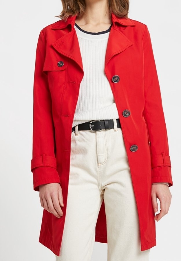 Abrigo trinchera rojo con cierre de botones, cuello con muescas y presillas de cinturón visibles, combinado con una camiseta blanca de canalé y pantalones beige.