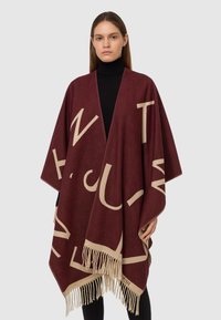 Poncho in lana bordeaux con grandi lettere beige, apertura frontale e orlo frangiato. Modella che indossa un dolcevita nero sotto.