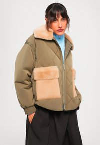 Donna che indossa una giacca trapuntata verde oliva con colletto di pelliccia beige e grandi tasche anteriori, camicia azzurra chiara e pantaloni larghi scuri, con le mani in tasca.
