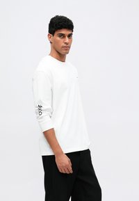 Hosszú ujjú fehér pamut ing kerek nyakkivágással és grafikai nyomattal a bal ujján. Fekete csíkozott nadrággal viselve.