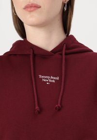 Felpa bordeaux con cappuccio e lacci, con logo bianco "Tommy Jeans New York". Tessuto di cotone morbido, vestibilità ampia e dettagli a coste.