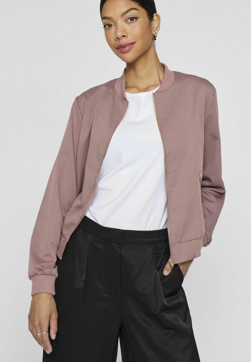 Bomberjacke in Mauve, mit einem goldenen Reißverschluss, gerippten Bündchen und einer lässigen Passform. Getragen über einem weißen T-Shirt und kombiniert mit schwarzen Hosen.