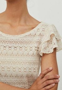 Top in pizzo crema con scollo ondulato e maniche corte a volant. Motivi floreali e geometrici su tutto il capo. Tessuto morbido e texturizzato.