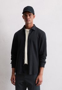 Young man wearing a black cap, black button-up shirt over a white t-shirt, and black pants standing against a plain white wall.