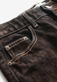 Bruna denimjeans har en bakficka med sydda detaljer och kopparnitar, vilket visar en strukturerad yta och ljus kontrastsömnad.