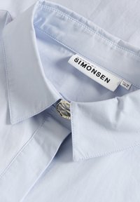 Lichtblauw overhemd met kraag en een gladde textuur, voorzien van een zilveren knoopaccent en een label met de tekst "KAREN SIMONSEN."