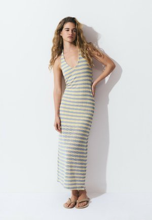 Striped maxi dress in light blue and yellow, halter neck design, textured fabric, with a side slit and paired with tan sandals.