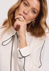 Biała bluzka z czarnym wykończeniem, z dekoltem w kształcie kokardy. Modelka nosi złotą i białą pierścionek oraz duże czarne kolczyki. Miękka faktura.